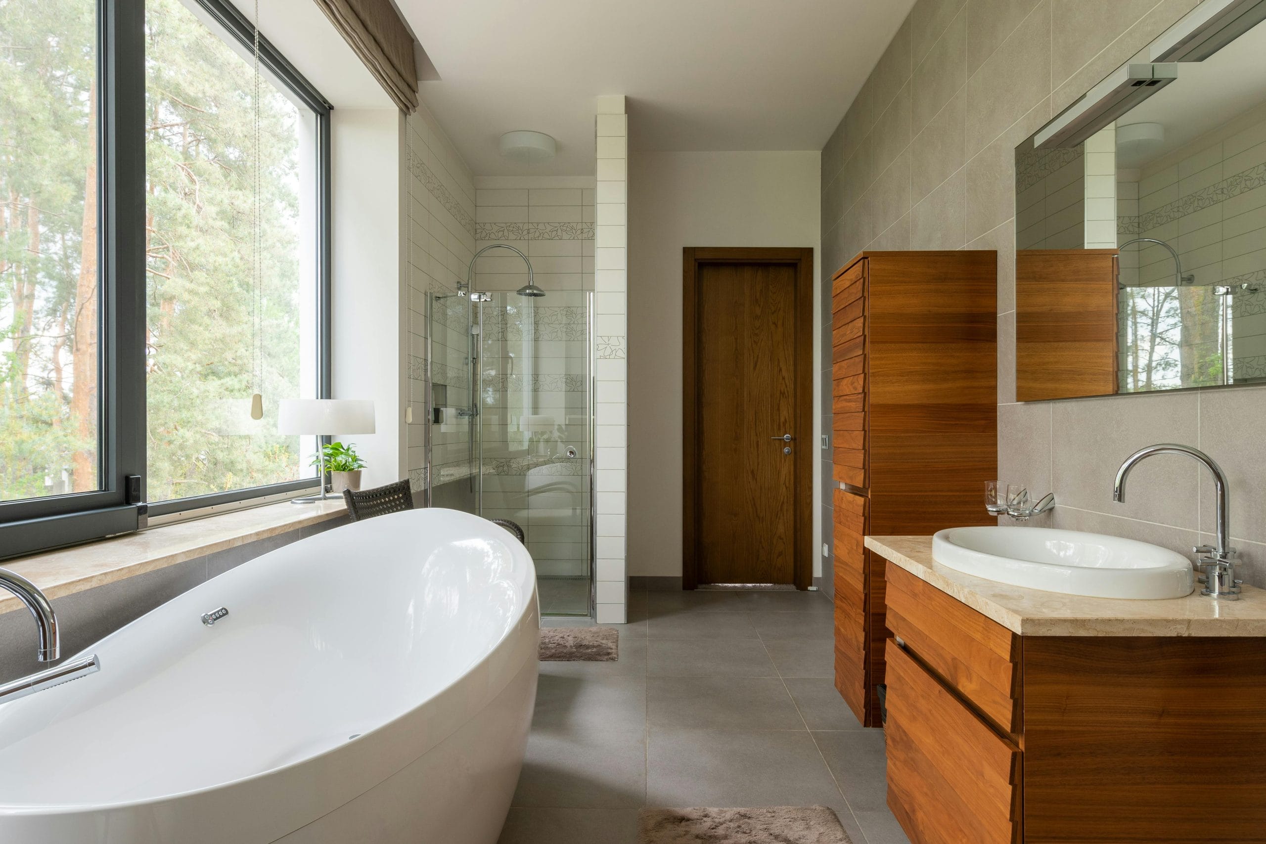 Modern bathroom featuring a freestanding white bathtub, wooden cabinetry, large windows with greenery view, and a glass shower enclosure, illustrating stylish home extension design ideas.